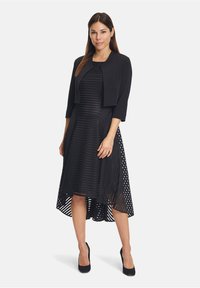 Musta mekko, jossa on tiukkanen bodice ja epäsymmetrinen helma, jossa on raidallinen rakenne ja pilkkudetaljit. Mukana lyhyt takki.