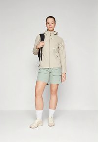 Beež veekindel jope kapuutsiga, kombineeritud mintroheliste cargo lühikeste pükstega. Modell kannab tossusid ja musta seljakotti, seisab neutraalsel taustal.