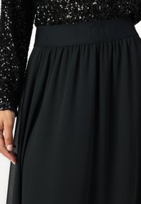 Čierna sukňa z mäkkej, splývavej látky s elastickým pásom, na ktorom je jemne embosované logo. Top so flitrami kontrastuje so sukňou.