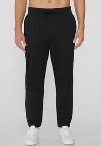 Pantalones de chándal negros con cintura elástica y cordón. Diseño con piernas ajustadas y puños acanalados. Llevados con zapatillas blancas.