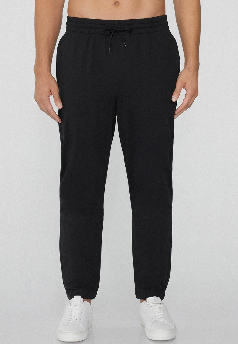 Pantalones de chándal negros con cintura elástica y cordón. Diseño con piernas ajustadas y puños acanalados. Llevados con zapatillas blancas.