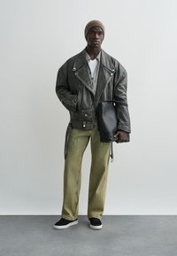 Nahast mootorratturite jakk, ruuduline särk, valge T-särk, oliivirohelised sametpükst, must kott, mustad tossud. Neutraalne taust, modell seisab sirgelt.