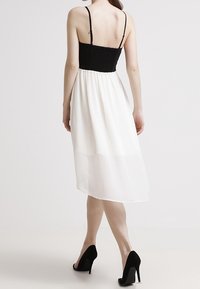 Zwarte gedrapeerde jurk met verstelbare bandjes en een vloeiende witte rok. De zoomlijn is asymmetrisch, en het model draagt zwarte hoge hakken.