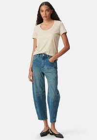 Vrouw in een beige blouse met korte mouwen, high-waisted blauwe jeans met knienaht, zwarte geweven slingback flats, staande met één hand in haar zak.