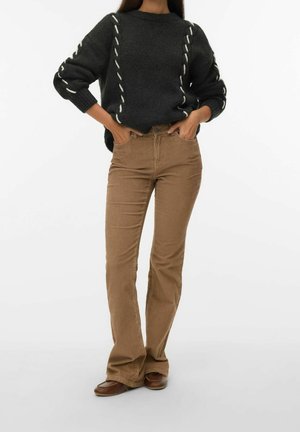 Femme portant un pull en tricot charbon avec des rayures blanches et un pantalon en velours côtelé beige taille haute avec des chaussures marron, les mains dans les poches.