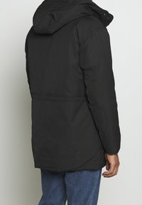 Czarna pikowana kurtka z kapturem, o gładkiej teksturze, widocznym przeszyciu i subtelnym logo na plecach.