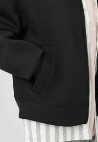 Sweatshirt noir zippé avec un tissu texturé, présentant une poche latérale et un ourlet côtelé, superposé à une chemise rayée.