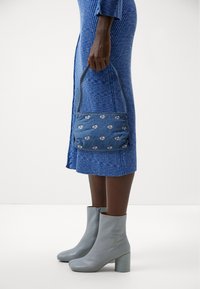 Modrá vzorovaná kabelka s texturovanou tkaninou, zdobená stříbrnými logy, doplněná světle šedými kotníkovými botami a žebrovanými modrými šaty.