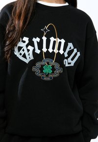Sudadera negra con texto blanco "Grimey" de gran tamaño. Presenta un trébol verde bordado y una cinta con las palabras en azul.