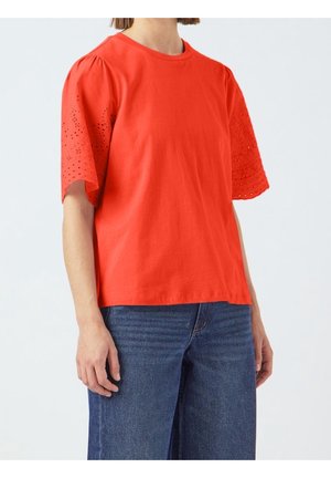 Osoba ubrana w jasnoczerwoną bluzkę z krótkim rękawem z haftem ażurowym na rękawach oraz niebieskie dżinsy, stojąca na białym tle.