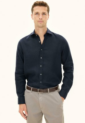 Mężczyzna w ciemnoniebieskiej koszuli zapiętej na guziki, włożonej do beżowych spodni z brązowym paskiem, stoi z jedną ręką w kieszeni.