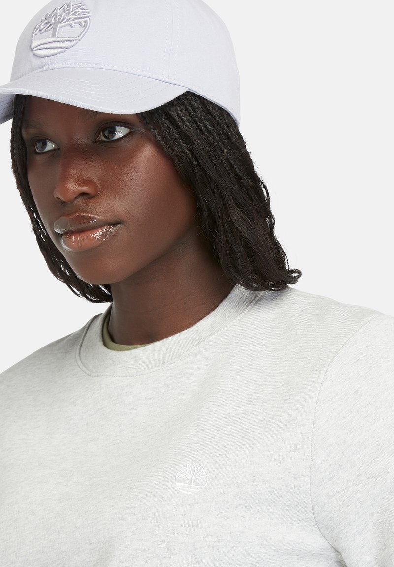 Felpa grigia chiara con scollatura rotonda e piccolo logo bianco. Il modello indossa un cappellino blu chiaro con un design semplice.