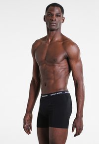 Fekete pamut boxer alsónadrág fehér derékrésszel, amelyen a "BJÖRN BORG" felirat látható, egy izmos alkattal rendelkező modellen bemutatva, sima háttér előtt.