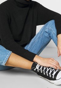 Une personne assise en tailleur portant un pull à col roulé noir, un jean bleu clair et des baskets montantes noires avec des lacets blancs.