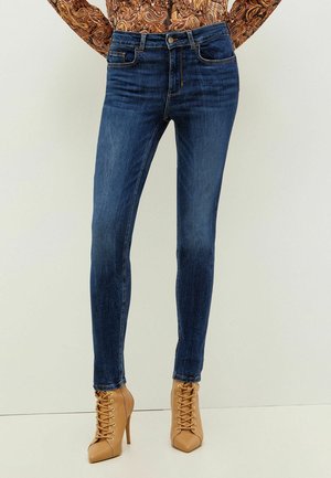 Blaue Slim-Fit-Jeans kombiniert mit beigen hochhackigen Schnür-Stiefeletten und einem braunen Paisley-Langarmtop.