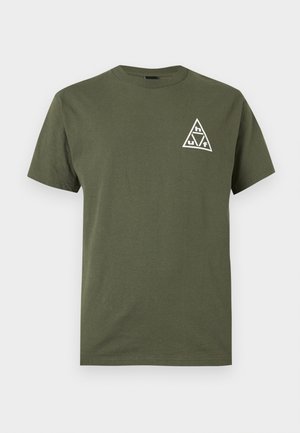 Mand iført olivengrøn T-shirt med stort hvidt trekantet logo med bogstaverne h, u og f på ryggen, stående mod en ensfarvet baggrund.
