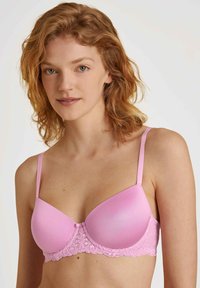 Lichtroze gewatteerde beha met kantaccenten, voorzien van verstelbare bandjes en een gladde bovenkant. Het kant voegt een gestructureerde detail toe onder de cups.