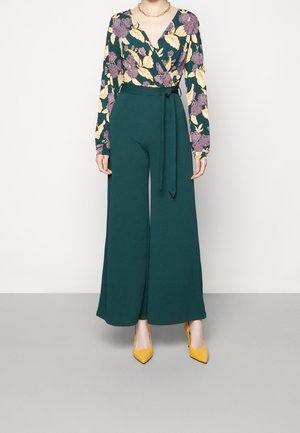 Femme portant un haut à manches longues à motif floral rentré dans un pantalon taille haute, jambes larges, vert foncé, avec ceinture nouée et des talons hauts jaunes.