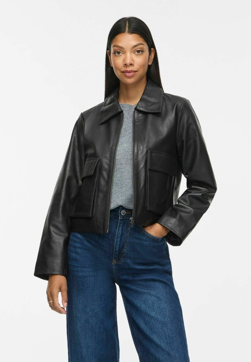 Femme aux longs cheveux foncés porte une veste en cuir noire par-dessus un pull gris et un jean bleu, debout avec une main dans la poche devant un fond blanc.