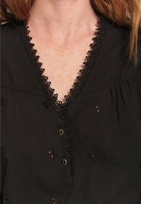 Blusa nera con ricami floreali, scollo a V con bordo in pizzo e bottoni dorati, realizzata in tessuto leggero con una vestibilità morbida.