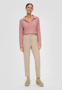 Jasnoróżowa koszula polo z długim rękawem i kołnierzykiem, zestawiona z beżowymi spodniami typu tapered. Modelka ma na sobie beżowe botki do kostki z teksturowaną podeszwą.