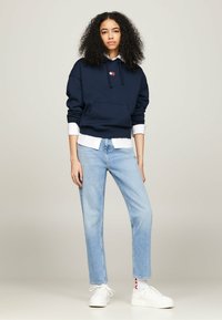 Mujer joven con cabello rizado, vistiendo una sudadera azul marino sobre una camisa blanca, jeans de mezclilla de color azul claro, zapatillas blancas y calcetines blancos con letras rojas.