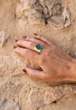 Hand mit dunkelrotem Nagellack, die einen goldenen Ring mit großem blauen ovalem Stein trägt und auf einer strukturierten beigen Steinfläche ruht.