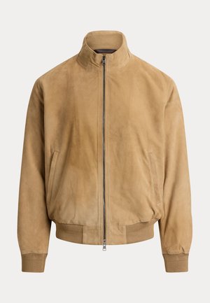 Homme portant une veste en daim beige, un pull en tricot vert clair, un pantalon blanc, des mocassins marron à pampilles et des lunettes de soleil foncées, debout devant un fond gris.