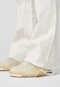 Baskets beiges avec une tige texturée, couture visible et unité d'air transparente ; associées à un pantalon large crème avec des ourlets effilochés.