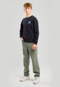 Mustapaitainen huppari, jossa on pieni logo rinnassa, yhdistettynä vihreisiin housuihin ja valkoisiin lenkkikenkiin. Kangas näyttää pehmeältä ja rentolta.