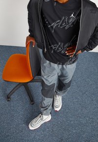 Personne portant une veste zippée noire, un t-shirt graphique noir, un pantalon de jogging gris et des baskets blanches, debout à côté d'une chaise de bureau orange sur un tapis bleu.