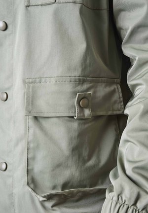 Chaqueta verde claro con una textura suave, botones a presión, un gran bolsillo delantero con solapa y puños fruncidos.