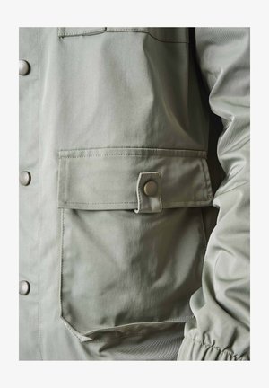 Veste vert clair avec une texture lisse, boutons pression, une grande poche avant avec rabat et des poignets froncés.