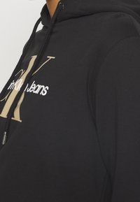 Svart hoodie i mjukt material, med en stor guldlogotyp och vit text "in KL Jeans." Inkluderar svarta dragsko och metalslitar.