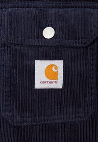 Marine corduroy stof met verticale ribbels, met een zak en een wit label met een oranje logo en de tekst "carhartt." Zilveren drukknoopdetail.