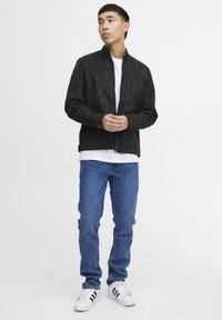 Jachetă bomber din suede negru, cu fermoar în față, buzunare laterale și guler structurat, asortată cu blugi albaștri și adidași albi.