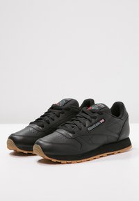 Svarte lær sneakers med tekstur, med en beige gummisåle, snøresystem og Reebok-logo på siden.