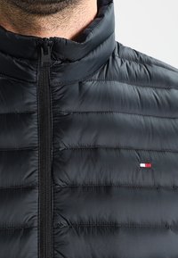 Black padded jacket with a high collar, featuring a full zipper and horizontal seam detailing. Small logo accent in red, white, and blue.