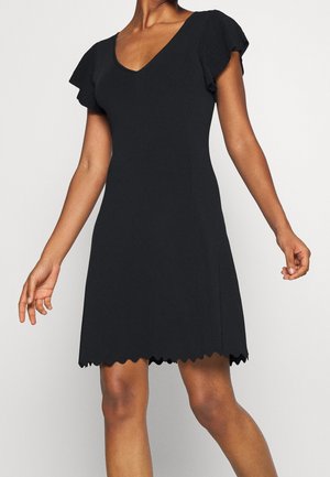 Kvinde iført sort kortærmet kjole med V-udskæring og bølget kant, stående foran en ensfarvet lys baggrund.