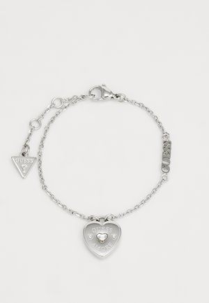 Bracciale in argento a catena con un ciondolo a forma di cuore con incisione "GUESS AMORE" e un piccolo accento a forma di cuore, insieme a un'etichetta triangolare.