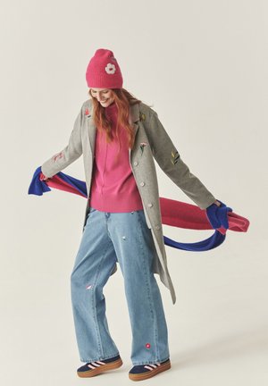 Hallloomine villane mantel lilleline tikand, roosa kootud beanie, roosa sviiter, laiad sinised teksad ja mitmevärviline sall. Triipudega tossud.