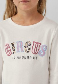 Junges Mädchen trägt ein weißes Shirt mit dem Aufdruck "Zirkus ist um mich herum" in bunten, gemusterten Buchstaben auf der Vorderseite.