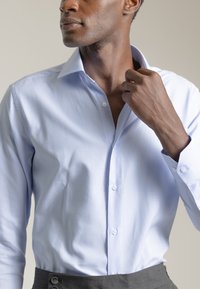 Camisa de dress azul claro con un patrón sutil, tapeta de botones y cuello tipo spread. La tela parece suave y nítida.