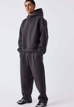 Jonge man staand, gekleed in een losse donkergrijze hoodie en bijpassende wijde broek met zwart-witte sneakers op een effen lichte achtergrond.
