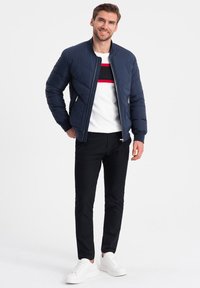 Námořnická proševaná bomber bunda s vroubkovaným límcem, zipovým zapínáním, bočními kapsami; nošena přes bílou svetr s červenými a černými pruhy a černými kalhotami, bílé tenisky.