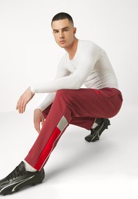 Burgundy Jogginghose mit grauen und roten Seitenschlaufen, aus weichem Stoff, mit elastischem Bund; kombiniert mit schwarzen Footballschuhen und einem taillierten weißen Langarmshirt.