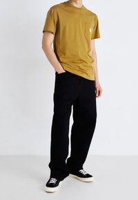 T-shirt en coton jaune moutarde avec poche et col rond, associé à un pantalon large noir et des baskets noires avec semelles blanches.