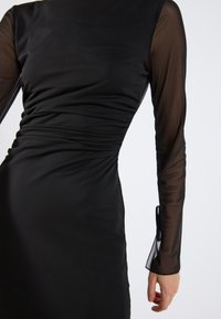 Abito nero con corpetto aderente e arricciature, maniche lunghe trasparenti e gonna dritta. Tessuto liscio con una leggera lucentezza.