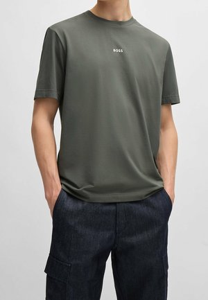 Homme portant un T-shirt à manches courtes vert olive avec le texte « BOSS » sur la poitrine et un pantalon cargo bleu foncé, les mains dans les poches.