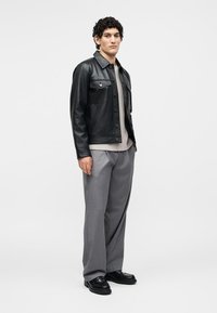 Νέος άνδρας που φοράει μαύρο δερμάτινο μπουφάν, μπεζ πουλόβερ, γκρι παντελόνι και μαύρα παπούτσια σε απλό λευκό φόντο.